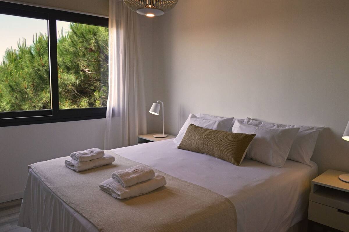A hotel room with a bed, two bedside lamps, a window with curtains, and a view of trees.