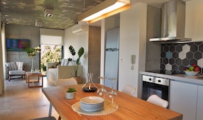 A modern kitchen with a built-in oven, a dining area with a wooden table and chairs, and a living area with a sofa and plants.