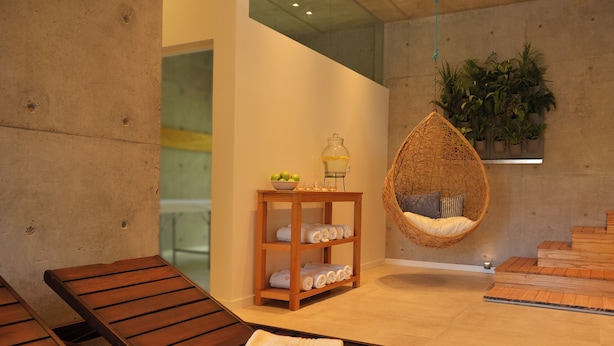 Un cuarto de spa con camastros de madera, una silla colgante de ratán, una mesita con dispensador de bebidas y un estante para plantas en la pared.