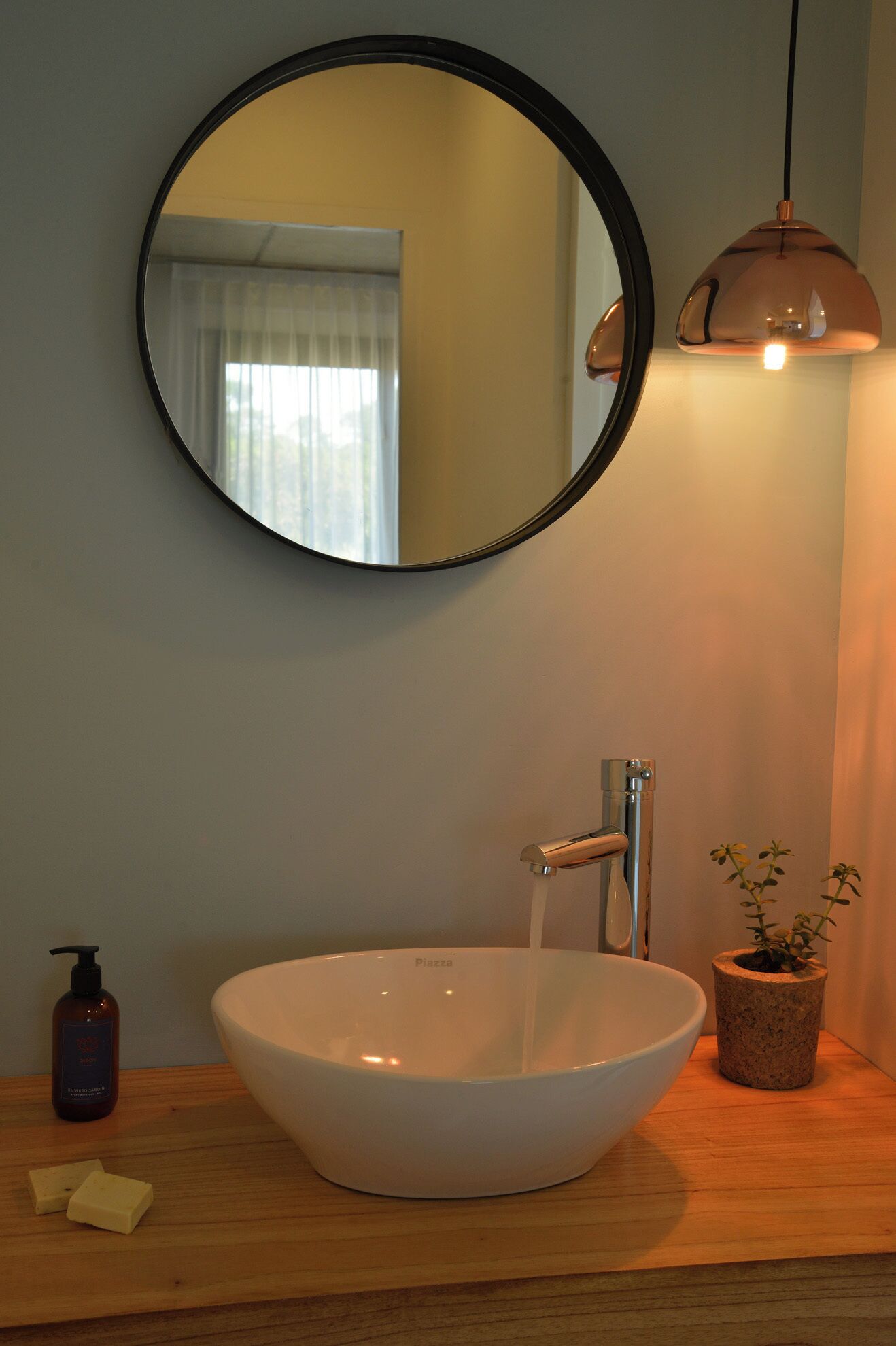 Une salle de bain avec un miroir rond, un lavabo sur colonne blanc, une douchette à main, un flacon de savon et une plante en pot.
