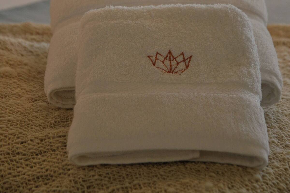 Two neatly folded white towels with a red embroidered symbol on a textured surface, a bed with white bedding, and a lamp in the background.