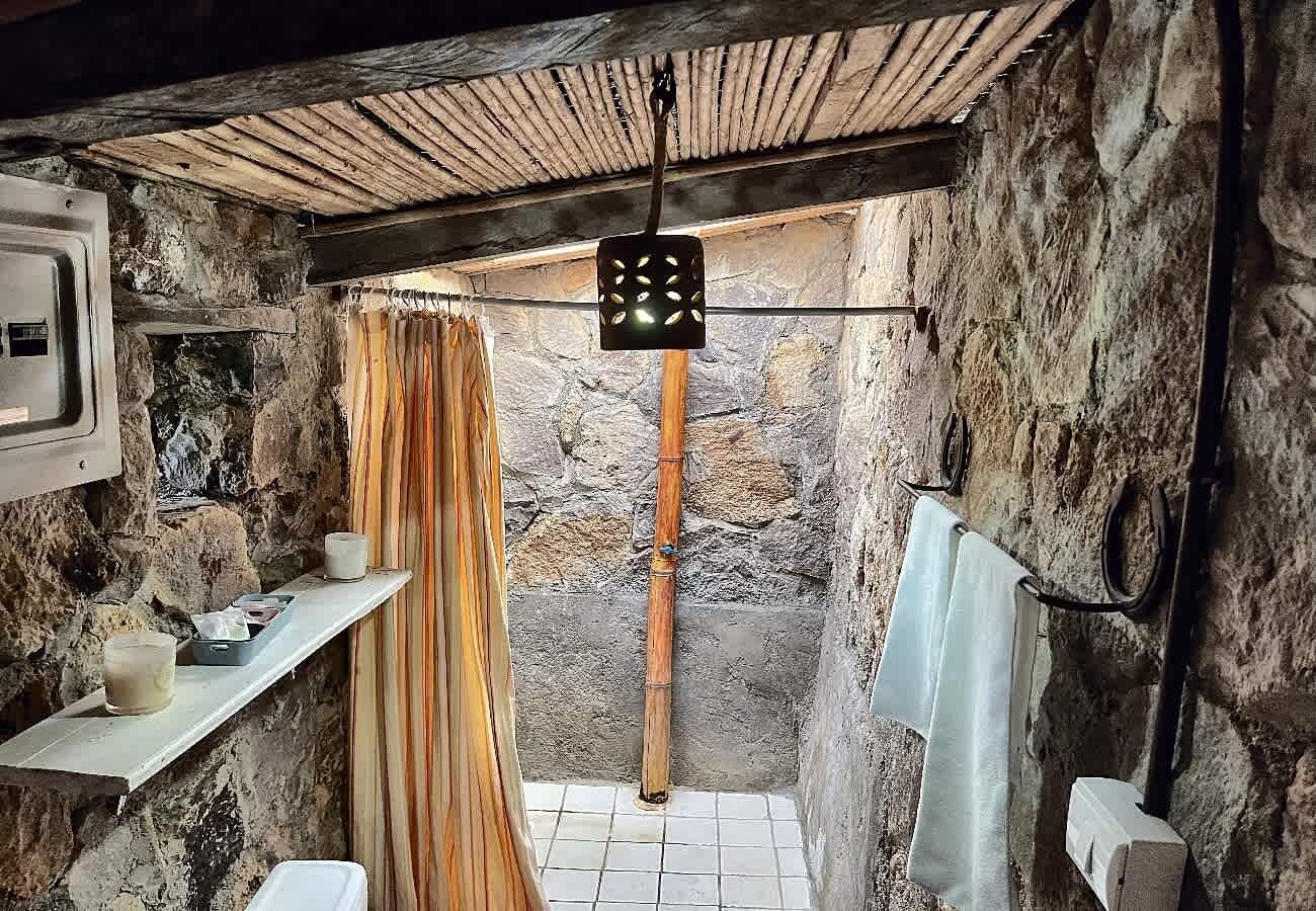 A rustic bathroom with a stone wall, a wooden shelf with candles, a bamboo shower curtain, and a hanging light fixture.