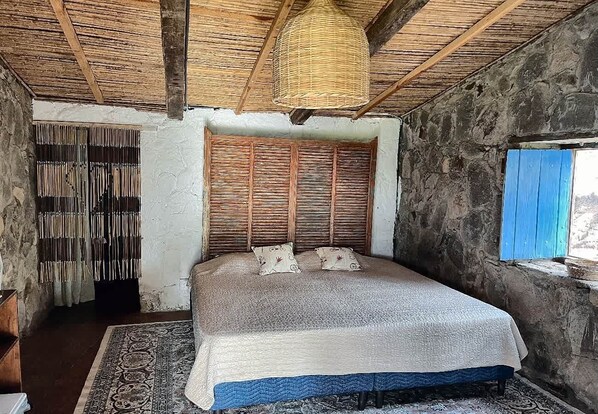 A bedroom with a bed, a woven headboard, a patterned rug, and a stone wall.