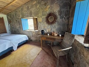 A rustic room with a bed, a table with a fan, a chair, and a decorative mirror.