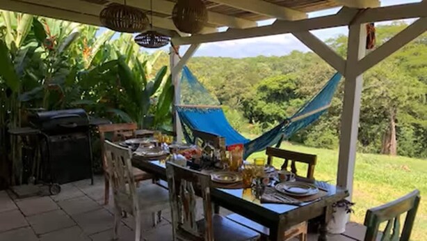 Un espace de restauration en plein air avec une table dressée pour deux personnes, entourée de chaises, d’un hamac et d’un barbecue.