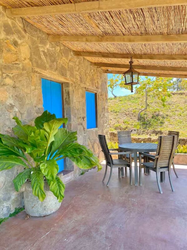 A stone building with a thatched roof, a table and chairs, a potted plant, and a blue door.
