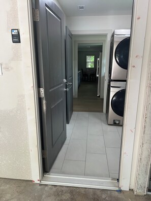 Un pasillo con una puerta cerrada, un área de lavandería con lavadoras apiladas y una vista de un comedor a lo lejos.