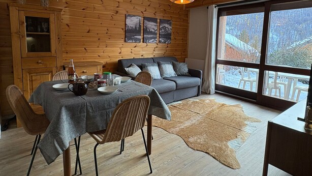 A cozy cabin interior with a dining table, chairs, a sofa, and a large window offering a view of snow-covered trees. - Comfortable studio with terrace and WIFI in Serre Chevalier (Serre Chevalier Chantemerle)