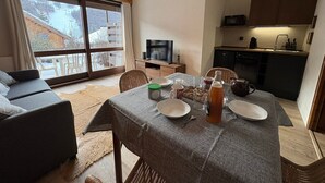 A cozy living room with a sofa, a dining table set with plates, a bottle, and a kettle. There's a television on a stand, a kitchen area with cabinets, and a large window with a view of a snowy landscape. - Comfortable studio with terrace and WIFI in Serre Chevalier (Serre Chevalier Chantemerle)