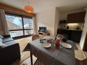 A cozy living room with a sofa, a dining table set with plates, a bottle, and a kettle. There's a television on a stand, a kitchen area with cabinets, and a large window with a view of a snowy landscape. - Comfortable studio with terrace and WIFI in Serre Chevalier (Serre Chevalier Chantemerle)