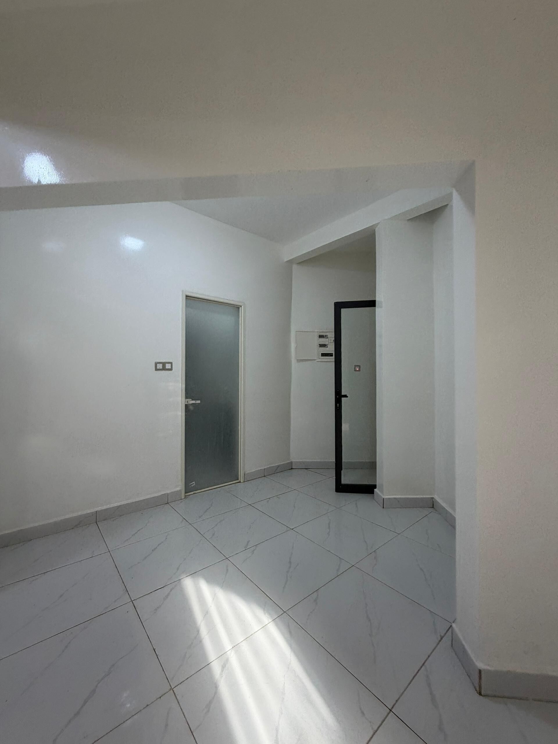 A spacious, empty room with white walls and tiled flooring, featuring two doors and a visible electrical panel.