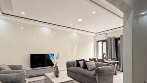 A modern living room with a flat-screen TV, grey sofas, a coffee table, and a large window. - Couramina (Dakar)