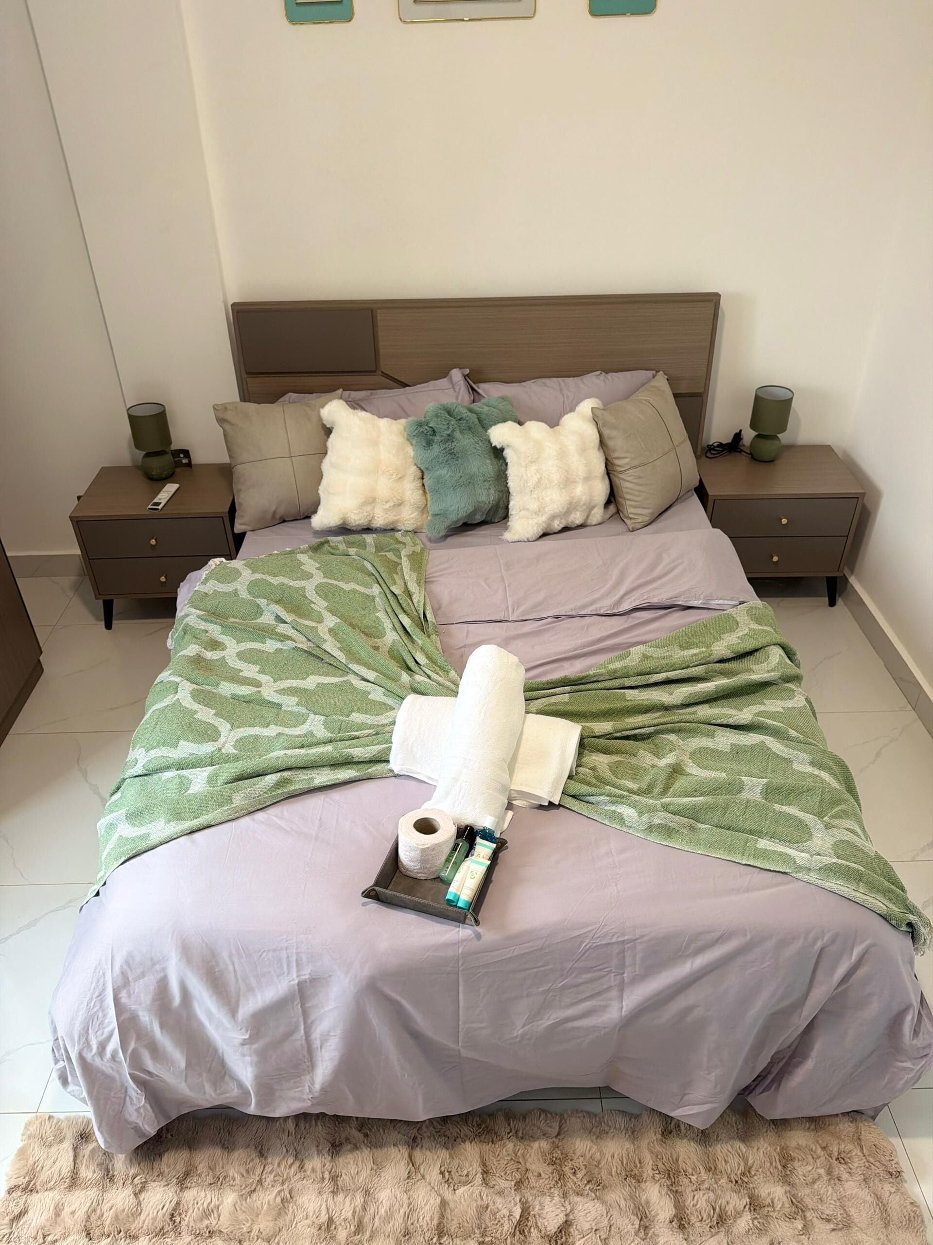 A neatly made bed with a green patterned blanket, a rolled-up towel, and toiletries on a tray. There are two bedside tables with lamps, and a fluffy rug on the floor.