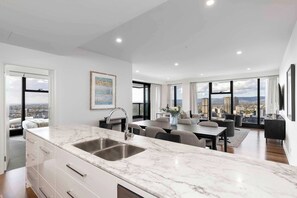 A modern kitchen with a marble countertop and a sink, a dining area with a table and chairs, and large windows offering a city view. - Luminesque Skyhome | Executive Residence in Adelaide CBD (Adelaide)
