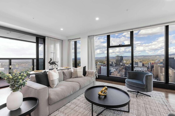 A modern living room with a large window, a sofa, a coffee table, and a chair. - Luminesque Skyhome | Executive Residence in Adelaide CBD (Adelaide)