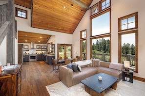 A spacious living area with a high ceiling, large windows, a kitchen, and a view of the outdoors. - The Cabin at Sundown. Hike All Day, Hot Tub All Night! (Bozeman)
