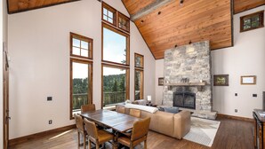 A spacious room with a large dining table, wooden chairs, a stone fireplace, and large windows offering a view of the outdoors.