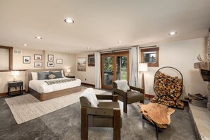 A bedroom with a bed, two chairs, a wooden table, a fireplace, and a large window.
