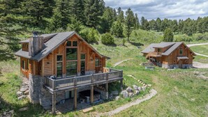 A wooden cabin with a balcony, stone foundation, and a surrounding grassy area with Adirondack chairs.