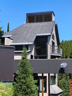 A modern multi-story building with a sloped roof, large windows, and a balcony. - Between the Pines | Ski, Hike, Fish, Soak, Repeat (Big Sky)