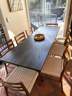 A dining table with a dark wooden surface and woven chairs, set on a tiled patio with a view of a snowy landscape through a glass door. - Between the Pines | Ski, Hike, Fish, Soak, Repeat (Big Sky)