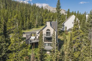 A modern two-story cabin with a hot tub and a deck, surrounded by dense forest and mountains.