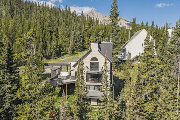 Eine moderne, zweistöckige Holzhütte mit Whirlpool und Terrasse, umgeben von dichtem Wald und Bergen.