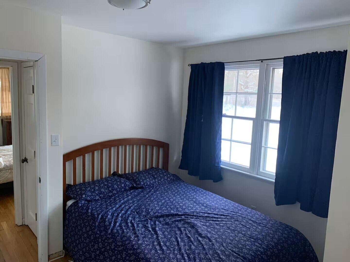 A bedroom with a bed, a window with curtains, a door, and a ceiling light.