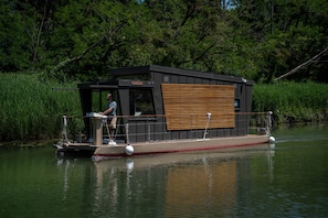 Une péniche dotée d’une terrasse, d’une petite cabine et d’une personne se tenant sur la terrasse.