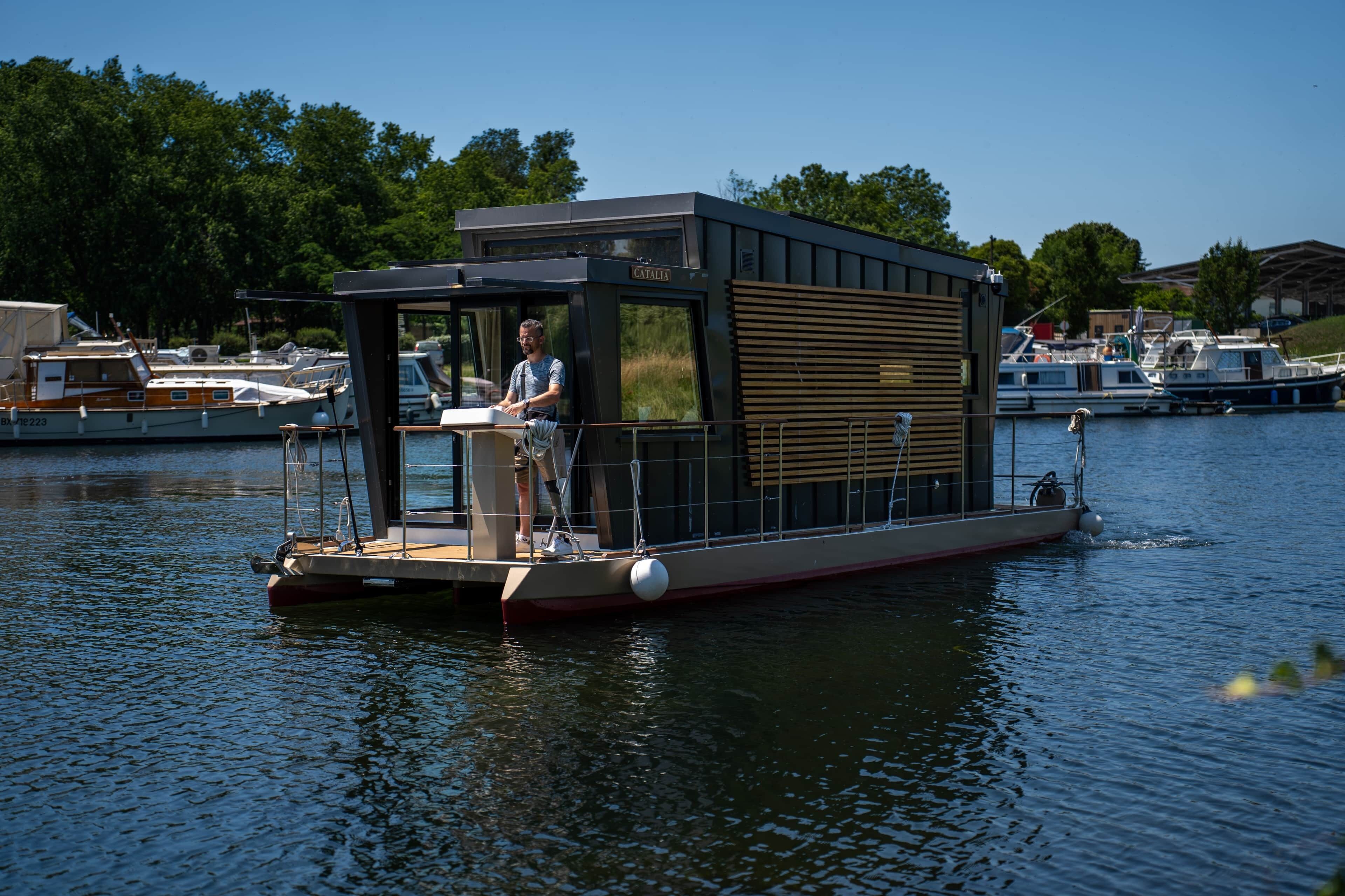 Une péniche dotée d’une petite terrasse, d’un espace abrité et d’une personne se tenant à la barre.