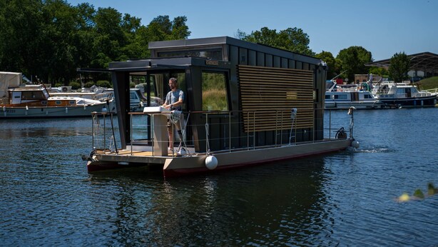 A houseboat with a small deck, a covered area, and a person standing at the helm.