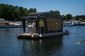 Una casa flotante con una pequeña cubierta, una zona techada y una persona al timón.