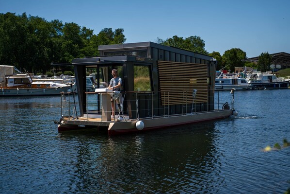 Une péniche dotée d’une petite terrasse, d’un espace abrité et d’une personne se tenant à la barre.
