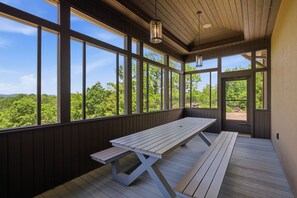 Ein überdachter Außenbereich mit einer Holzbank und einem Tisch, großen Fenstern und Blick ins Grüne.