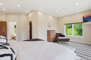 Ein Schlafzimmer mit einem großen Bett, einem Schminktisch, einem Sessel und einem Fenster mit Blick auf Grünflächen.