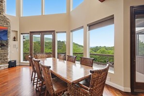 Un vaste espace dédié à la salle à manger, avec une grande table en bois et des chaises en osier, de grandes fenêtres offrant une vue sur des collines verdoyantes, et une cheminée en pierre.