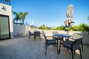 Eine Terrasse im Freien mit Tisch und Stühlen, einem schattenspendenden Sonnenschirm und einer Treppe zu einer erhöhten Ebene.