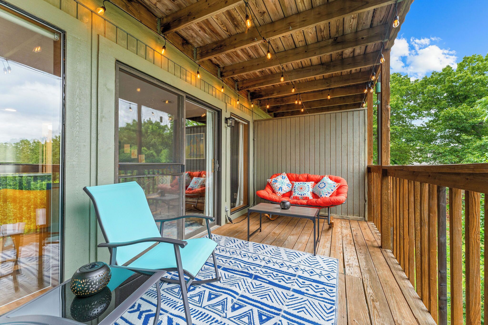 A covered outdoor area with a wooden deck, a glass door leading to a patio, and a seating area with a cushioned chair and table.