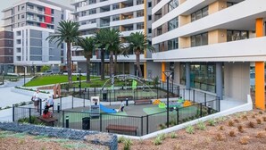 Un edificio moderno con diversi piani, un’area giochi con scivoli e altalene, e palme sullo sfondo.