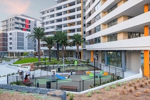 A modern building with multiple floors, a playground with slides and swings, and palm trees in the foreground. - A Little Slice of Paradise Pool, Gym+Parking (Macquarie Park)
