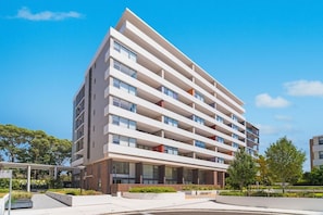 A multi-story building with a modern architectural design, featuring a flat roof, large windows, and a ground-level entrance. - A Little Slice of Paradise Pool, Gym+Parking (Macquarie Park)