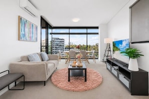 Una sala de estar moderna con un sofá, una mesa de centro, televisor y vistas a un paisaje urbano a través de amplios ventanales.