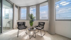 Een hoekkamer met twee zwarte stoelen, een bijzettafeltje, een potplant en grote ramen met jaloezieën.