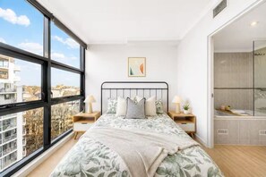 A bedroom with a large window, a bed with floral bedding, two bedside tables with lamps, a small plant, and a view of the city. - Surry Hills Chic 1BR in Prime Dining & City Spots (Surry Hills)