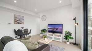 A modern living room with a flat-screen TV, a dining area with chairs, a sofa, and potted plants. - Designer 1BR Apt | Rooftop Oasis & Fast CBD Access (Rosebery)