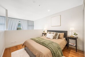 Une chambre à coucher avec un lit, deux tables de chevet avec des lampes, une fenêtre avec des rideaux et un tableau accroché au mur.