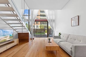 Un salon moderne avec une table basse en bois, un canapé gris et un téléviseur posé sur un meuble en bois. Un escalier mène à un balcon d’où l’on peut admirer les bâtiments et la verdure environnante.