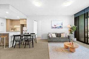 A modern living room with a grey sofa, a coffee table, and a dining area with a table and chairs. - Luxury 2BR Oasis – Panoramic CBD & Park Views (Sydney Olympic Park)