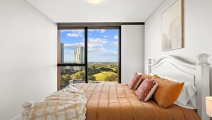 A bedroom with a large window, a bed with a patterned bedspread, and a view of buildings and greenery.