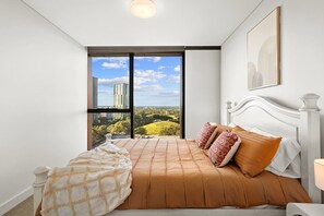 A bedroom with a large window, a bed with a patterned bedspread, and a view of buildings and greenery. - Luxury 2BR Oasis – Panoramic CBD & Park Views (Sydney Olympic Park)